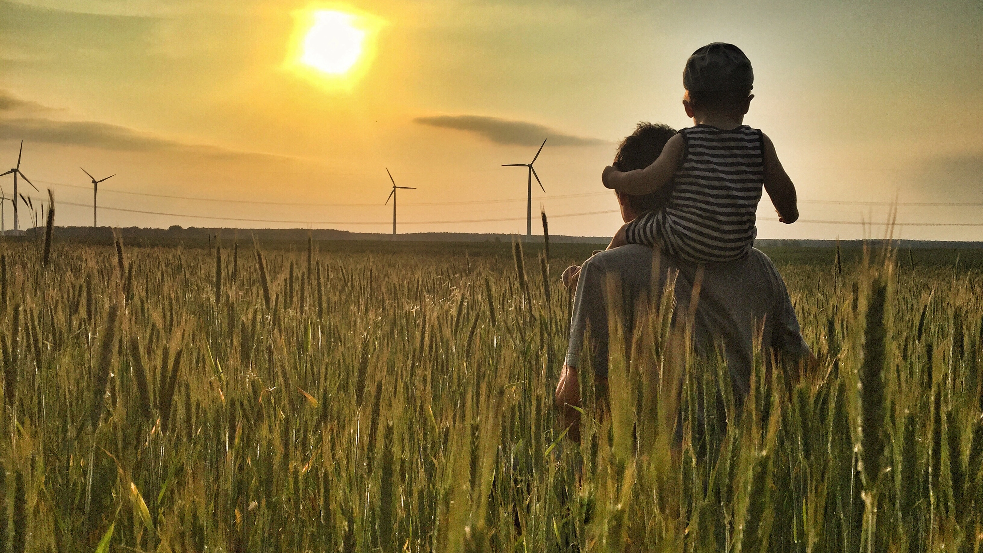 Man carries a child on his shoulders in a field at sunset with wind turbines in the background.
