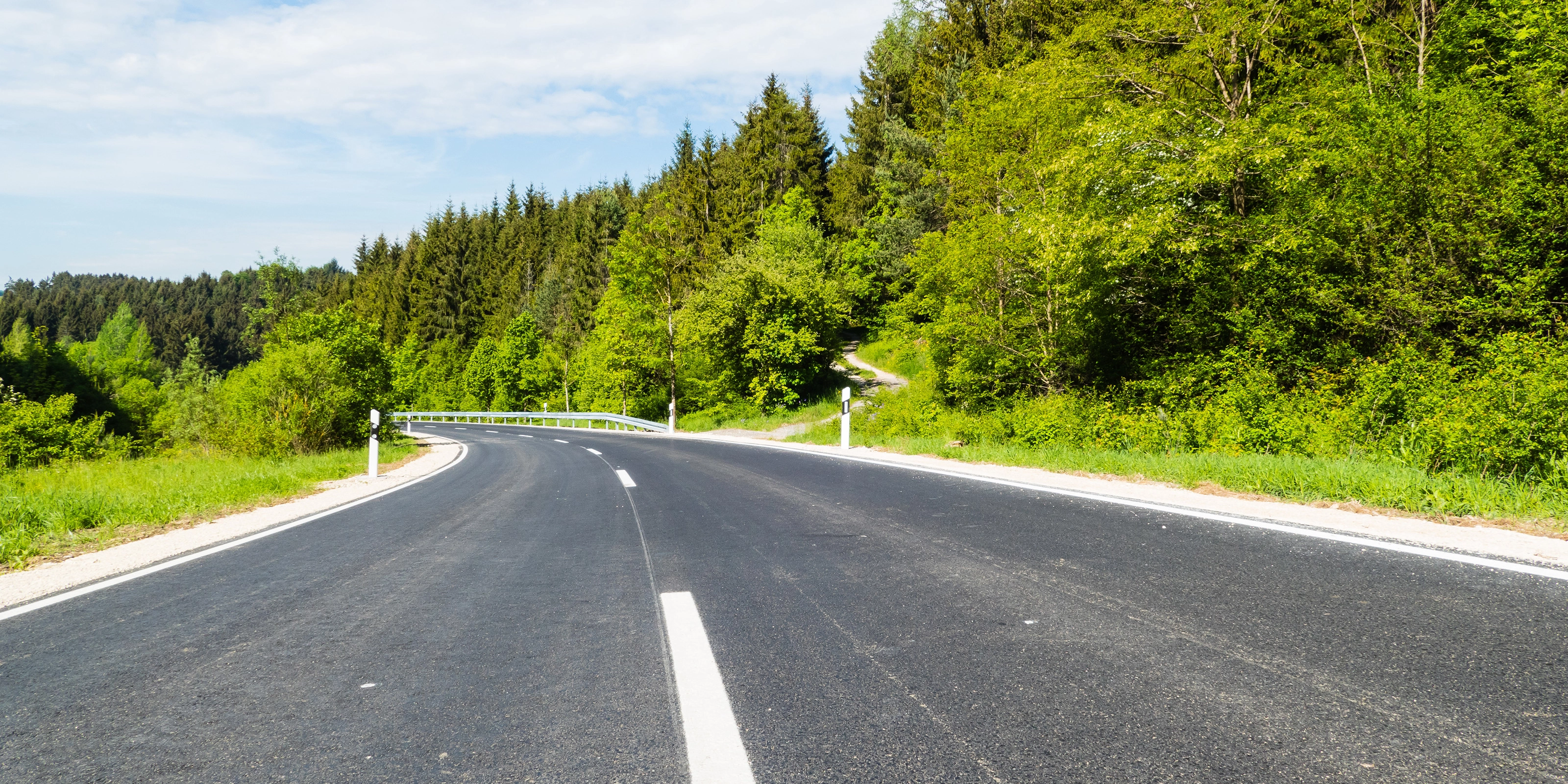 Empty asphalt road curves gently through a green forested landscape on a sunny day.