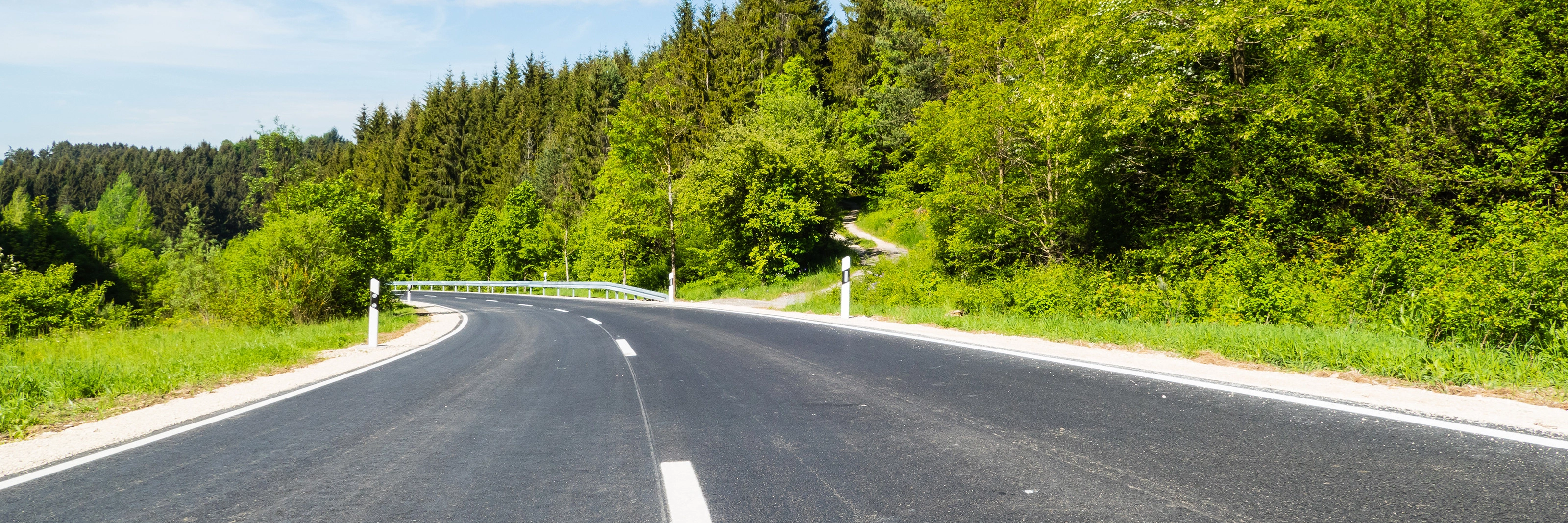 Empty asphalt road curves gently through a green forested landscape on a sunny day.