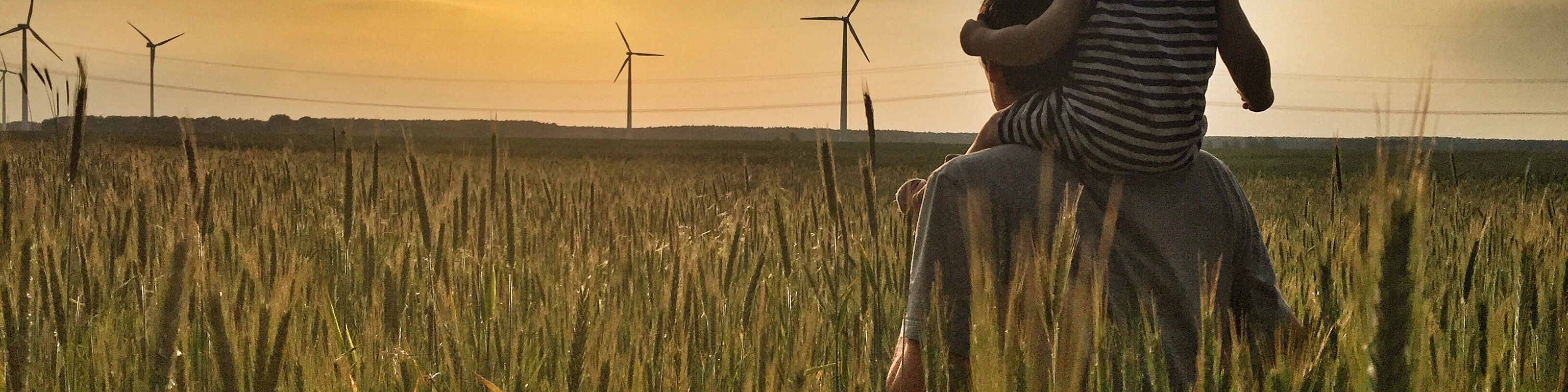 Man carries a child on his shoulders in a field at sunset with wind turbines in the background.