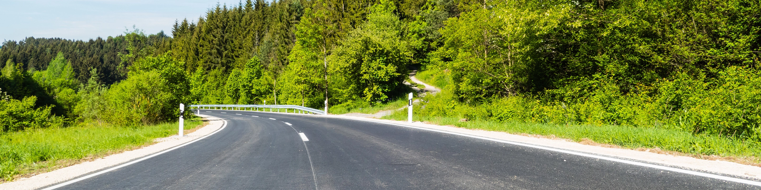 Empty asphalt road curves gently through a green forested landscape on a sunny day.