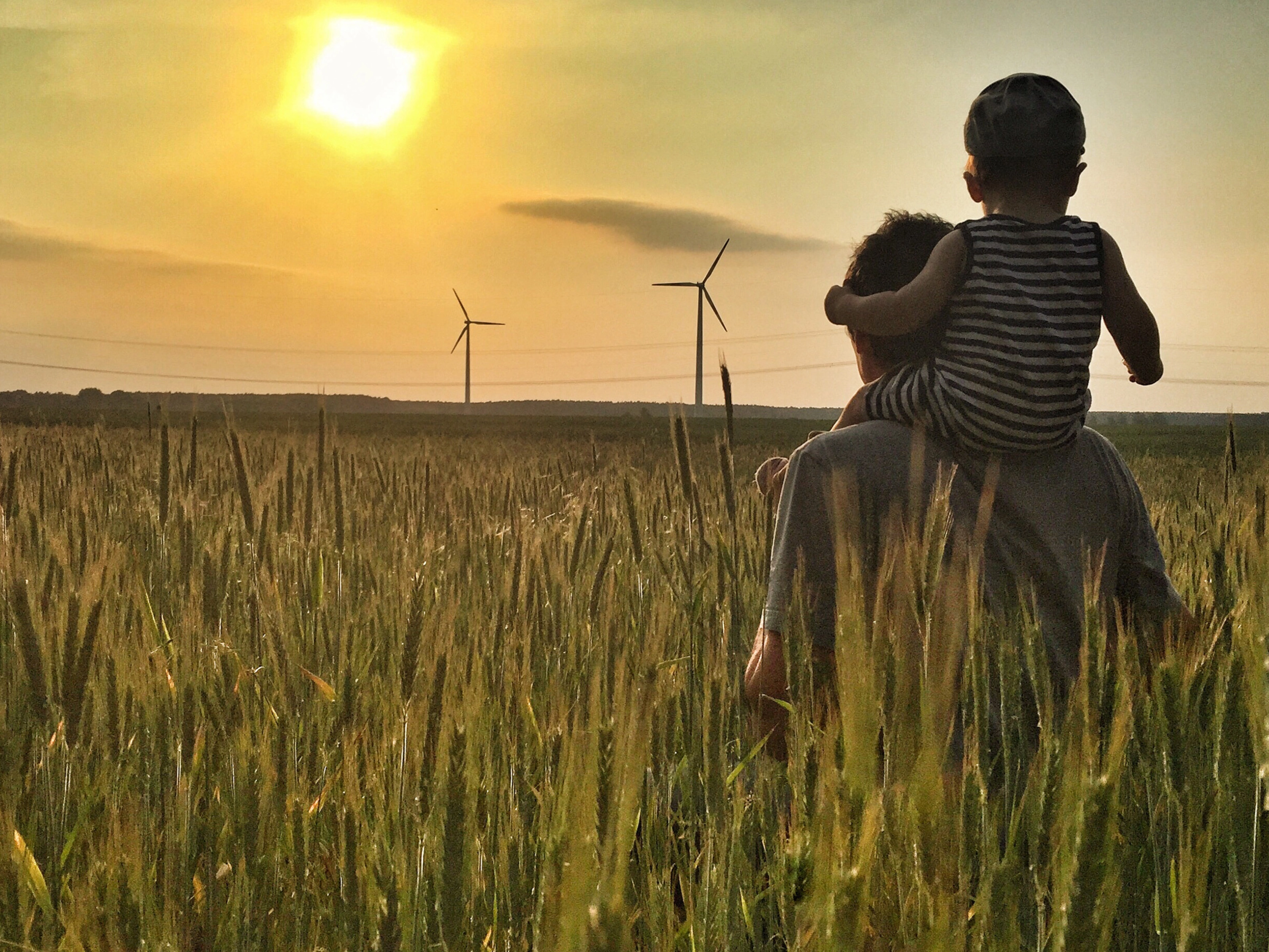 Man carries a child on his shoulders in a field at sunset with wind turbines in the background.
