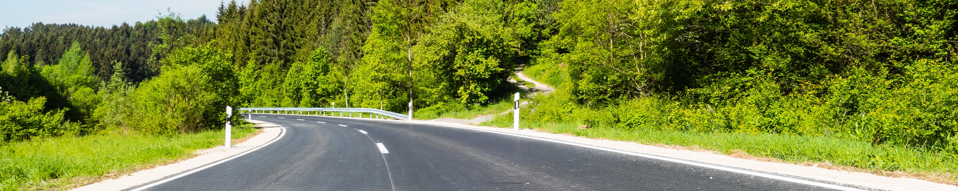 Empty asphalt road curves gently through a green forested landscape on a sunny day.