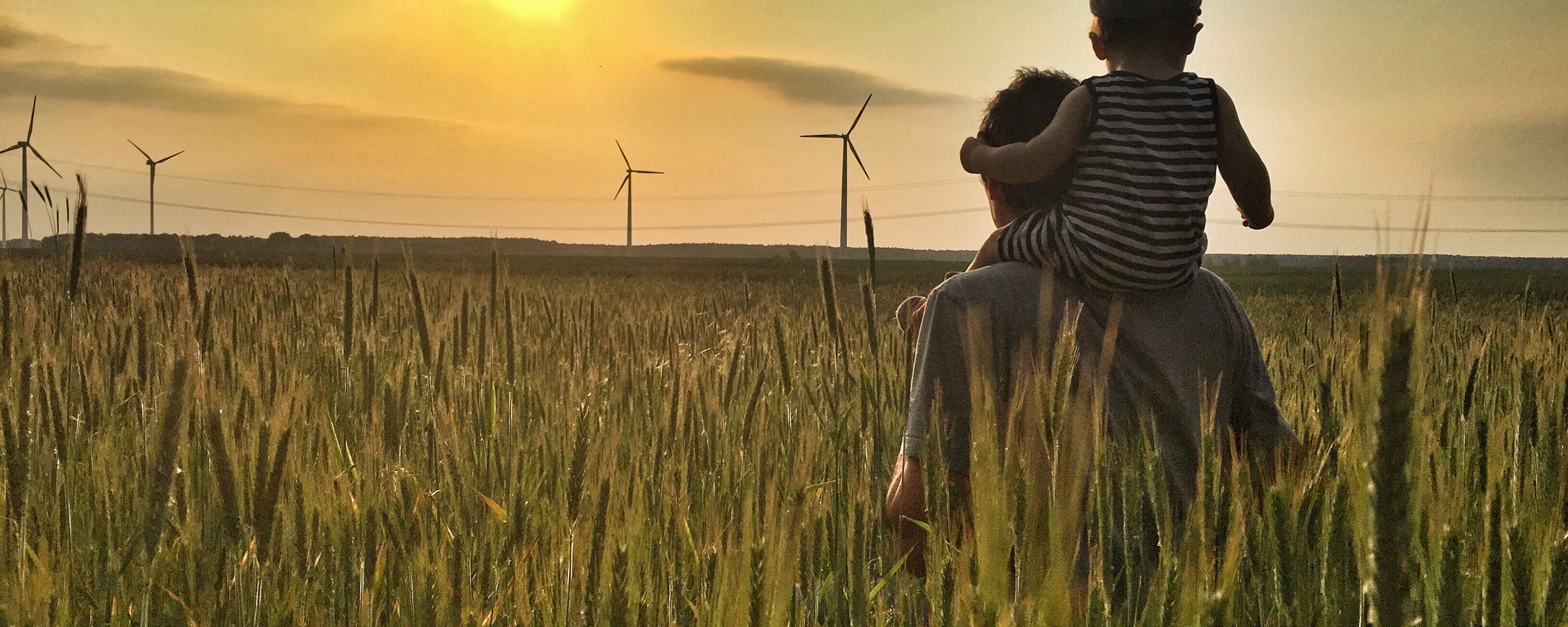Man carries a child on his shoulders in a field at sunset with wind turbines in the background.