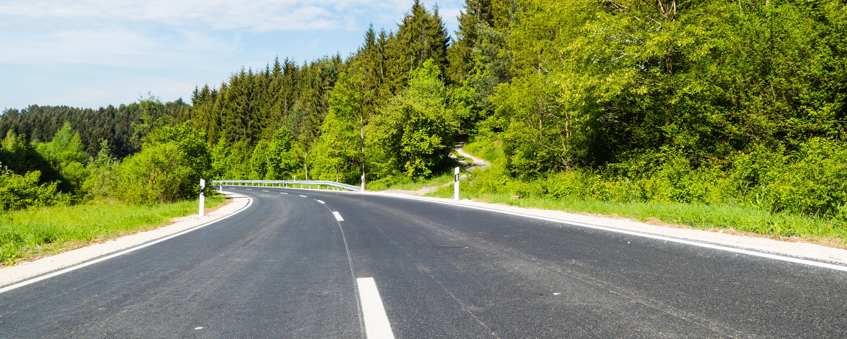 Empty asphalt road curves gently through a green forested landscape on a sunny day.