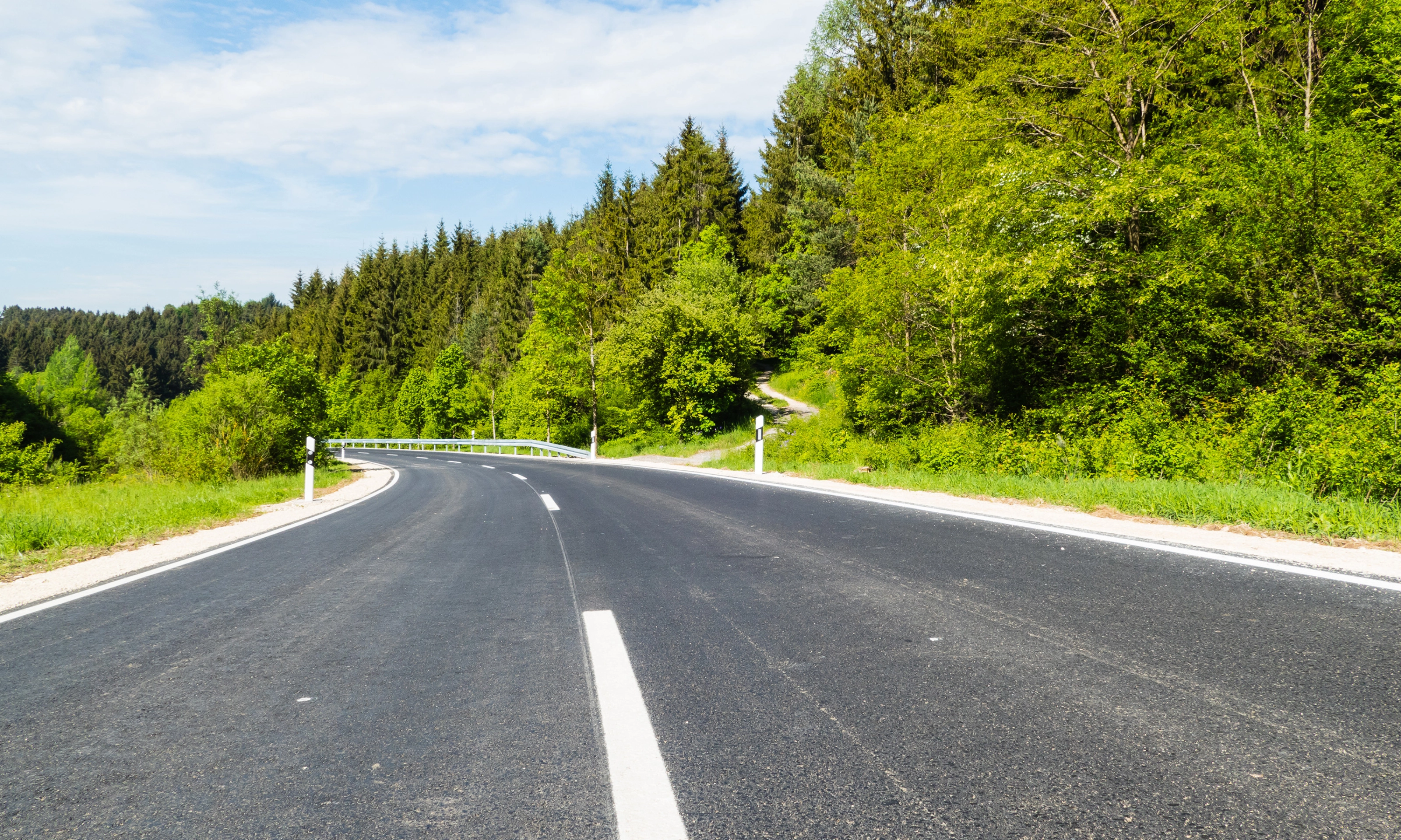Empty asphalt road curves gently through a green forested landscape on a sunny day.
