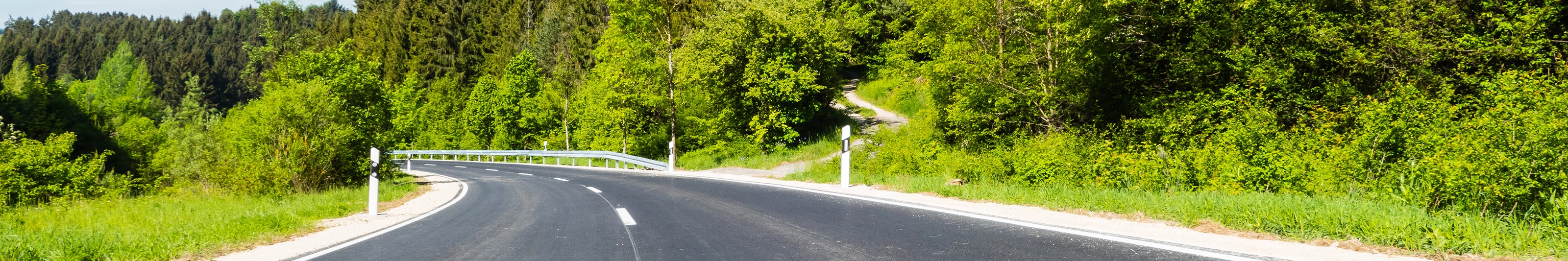 Empty asphalt road curves gently through a green forested landscape on a sunny day.