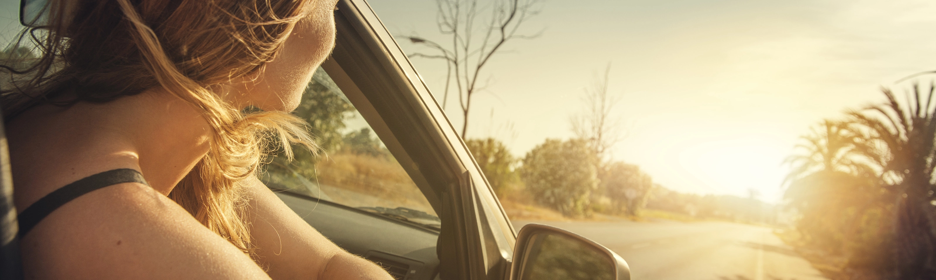 A woman leans out of a car window, looking at the sunset on a country road.