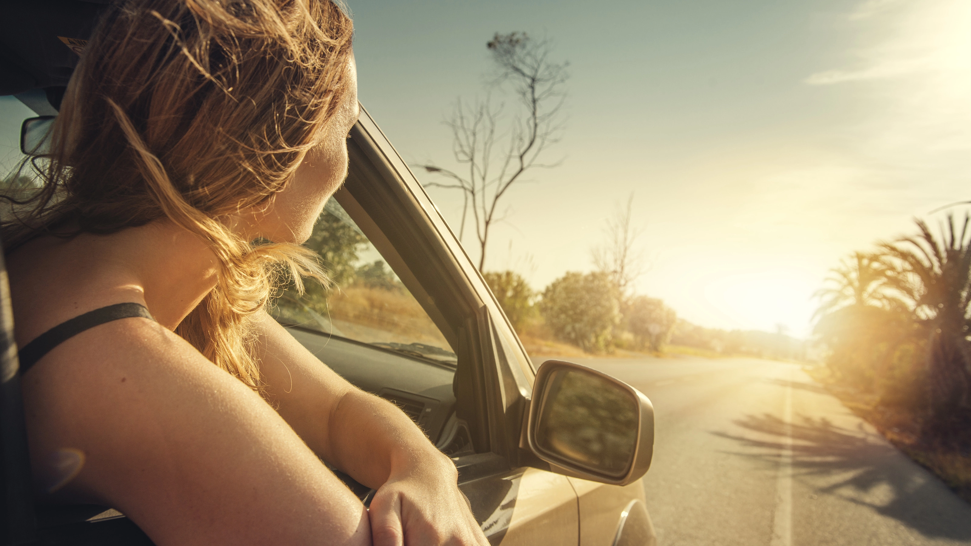 A woman leans out of a car window, looking at the sunset on a country road.