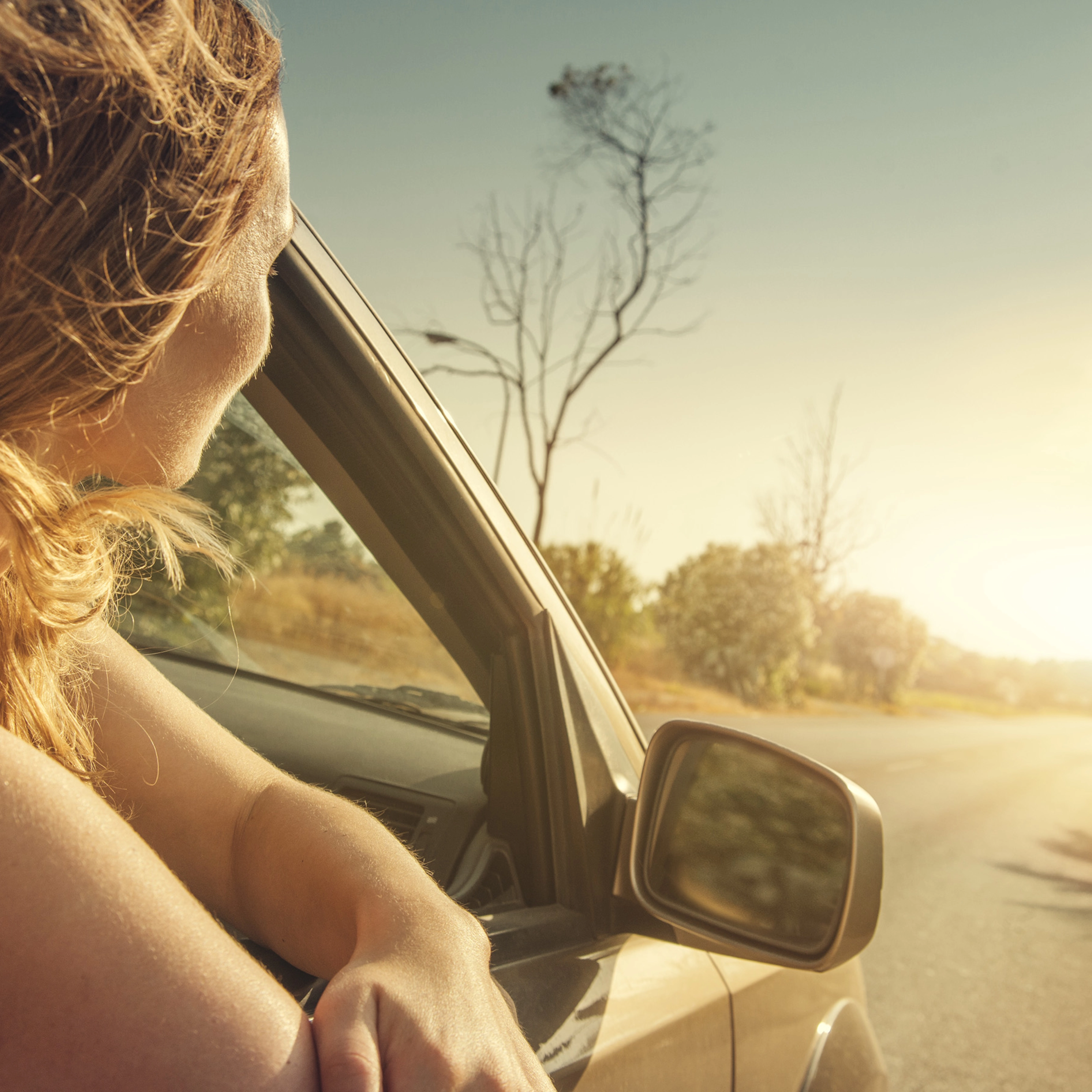 A woman leans out of a car window, looking at the sunset on a country road.