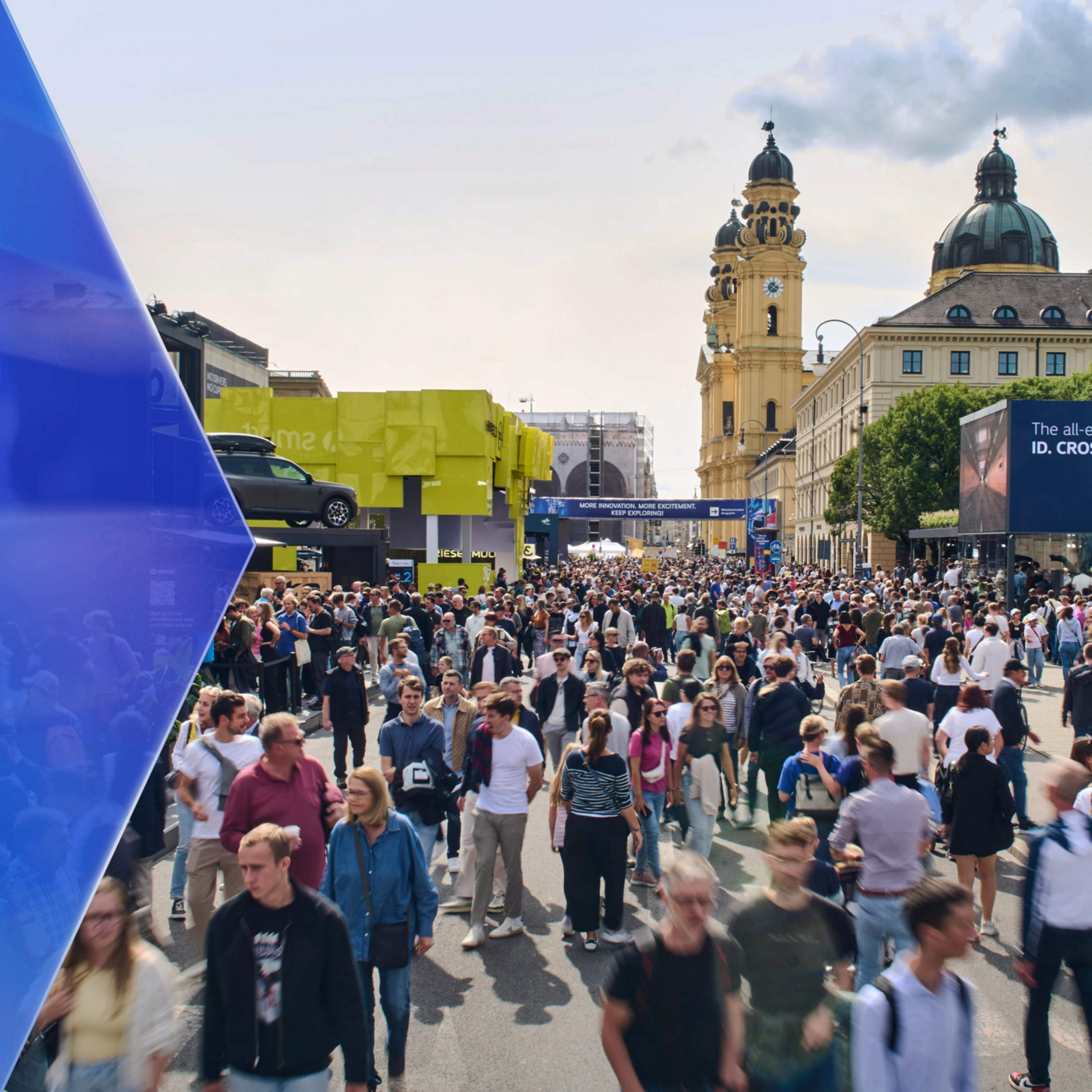 Large crowd at an outdoor mobility fair with exhibition booths and historic buildings in the background.
