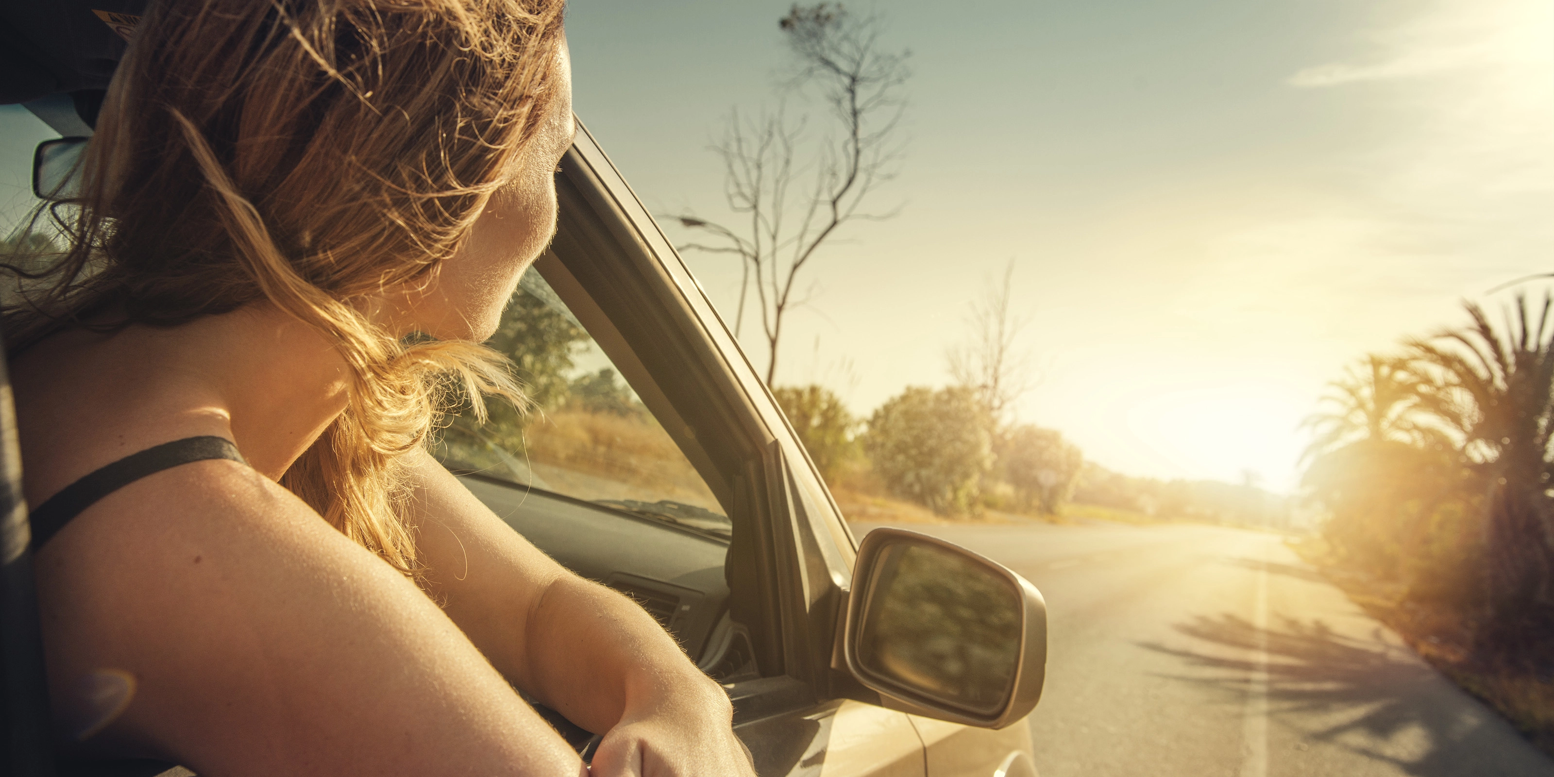 A woman leans out of a car window, looking at the sunset on a country road.