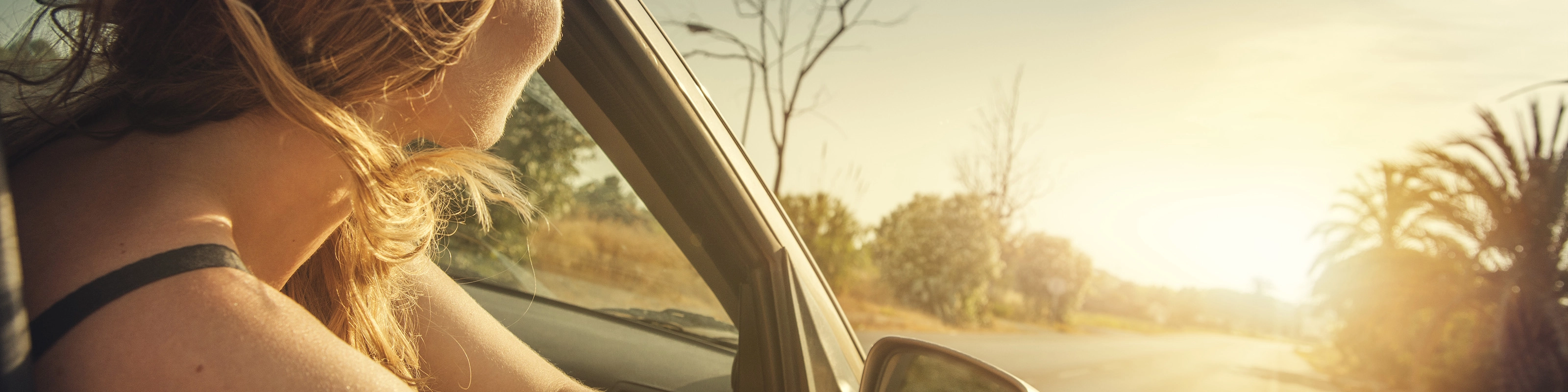 A woman leans out of a car window, looking at the sunset on a country road.