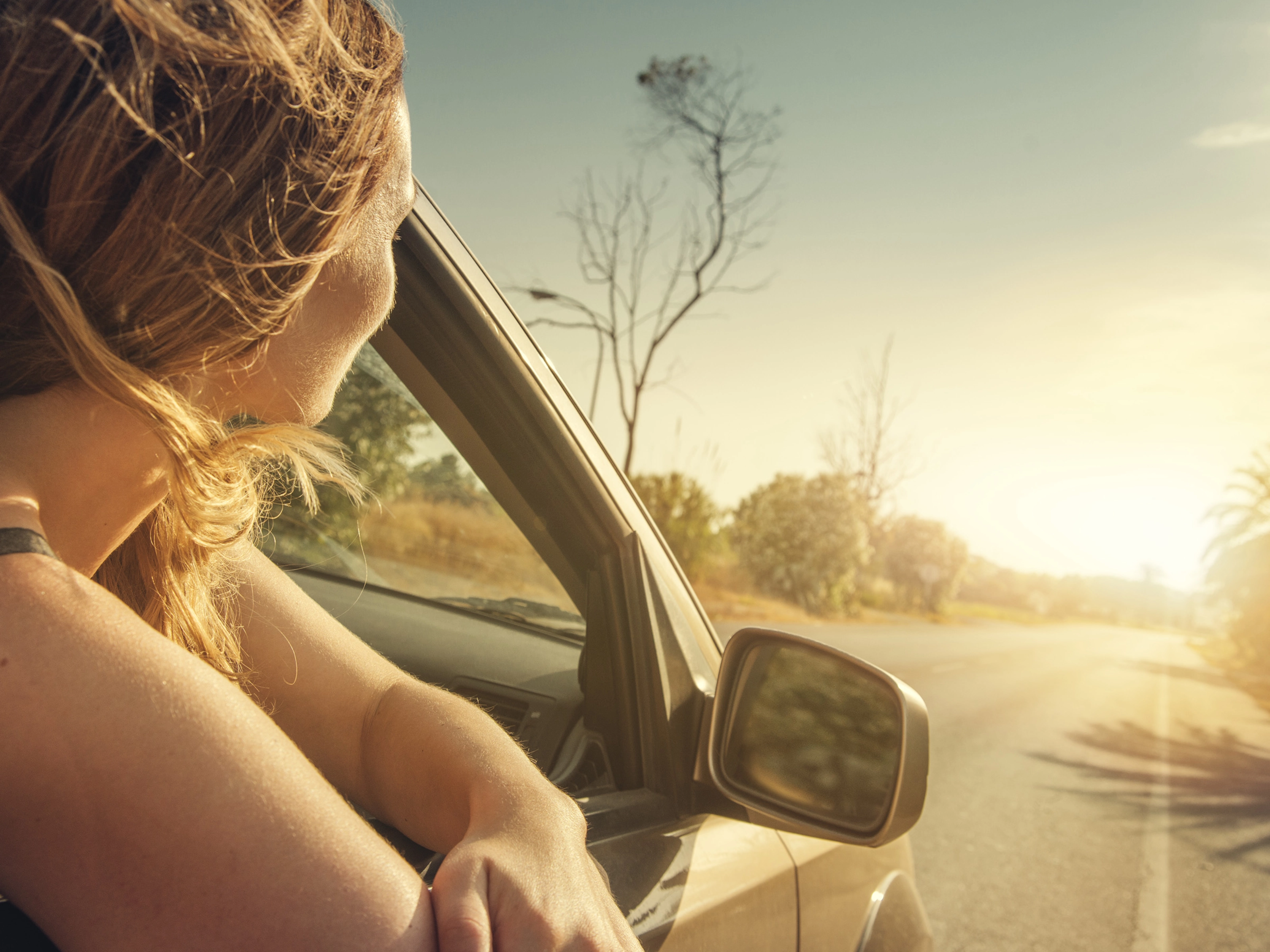 A woman leans out of a car window, looking at the sunset on a country road.