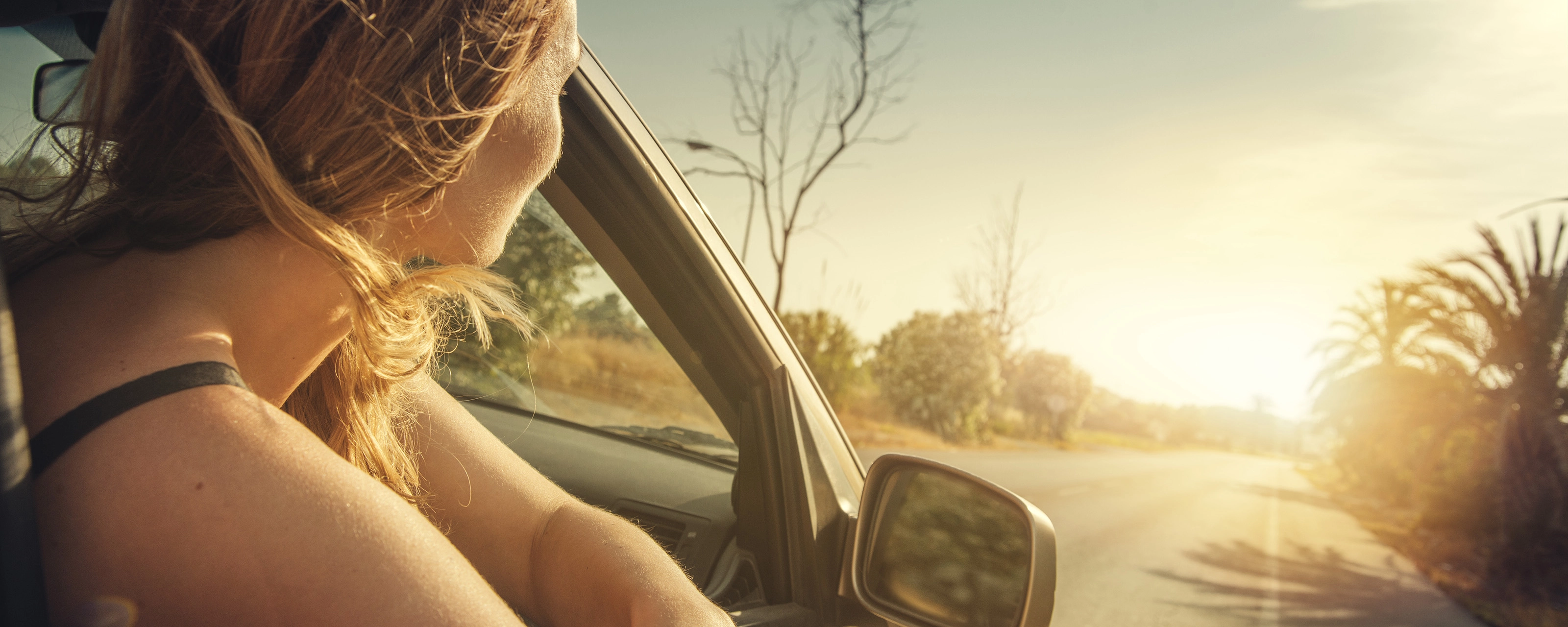 A woman leans out of a car window, looking at the sunset on a country road.